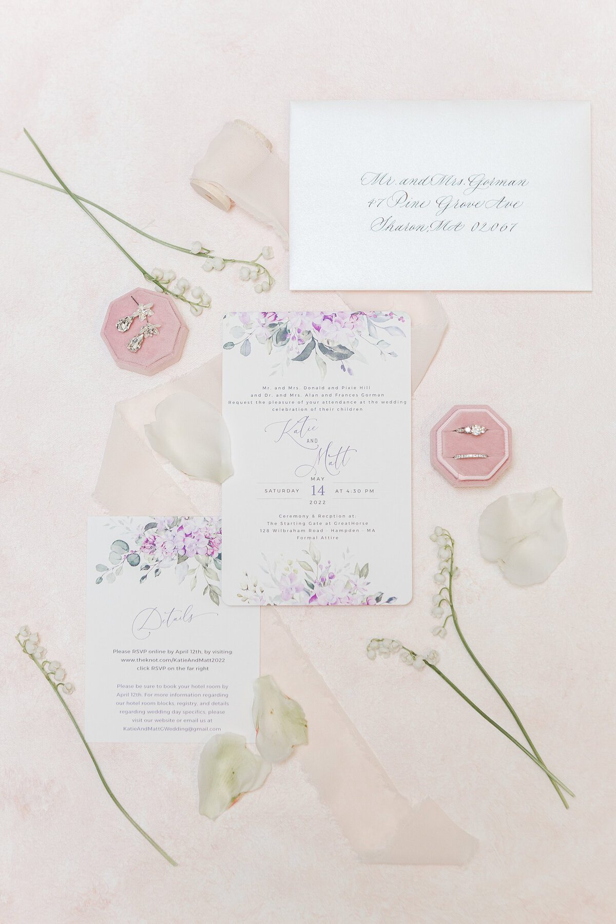 Wedding stationary is featured in a flat lay capturing the details. The image is accented with the rings, ribbon, flowers, and flower petals.