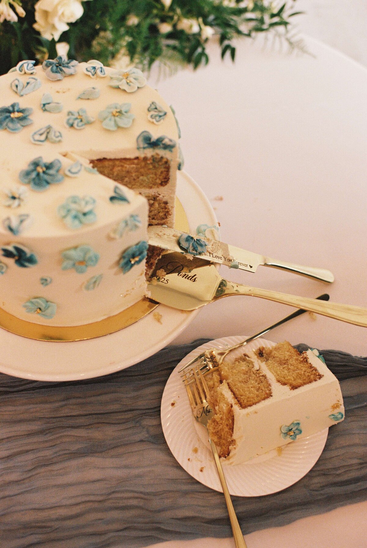 A slice of cake with white frosting and blue floral decorations sits next to a matching larger cake on a white plate. The setting feels elegant.