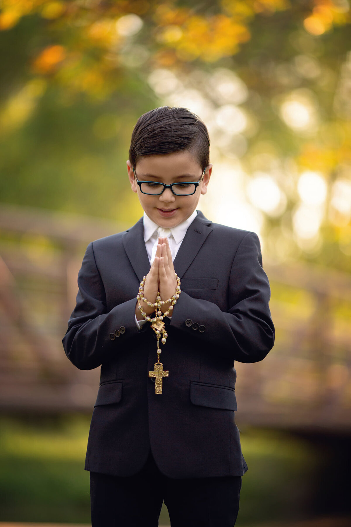 A young boy in a black suit prays in a park at sunset with gold rosary beads taken by a New Jersey Communion Portrait Photographer