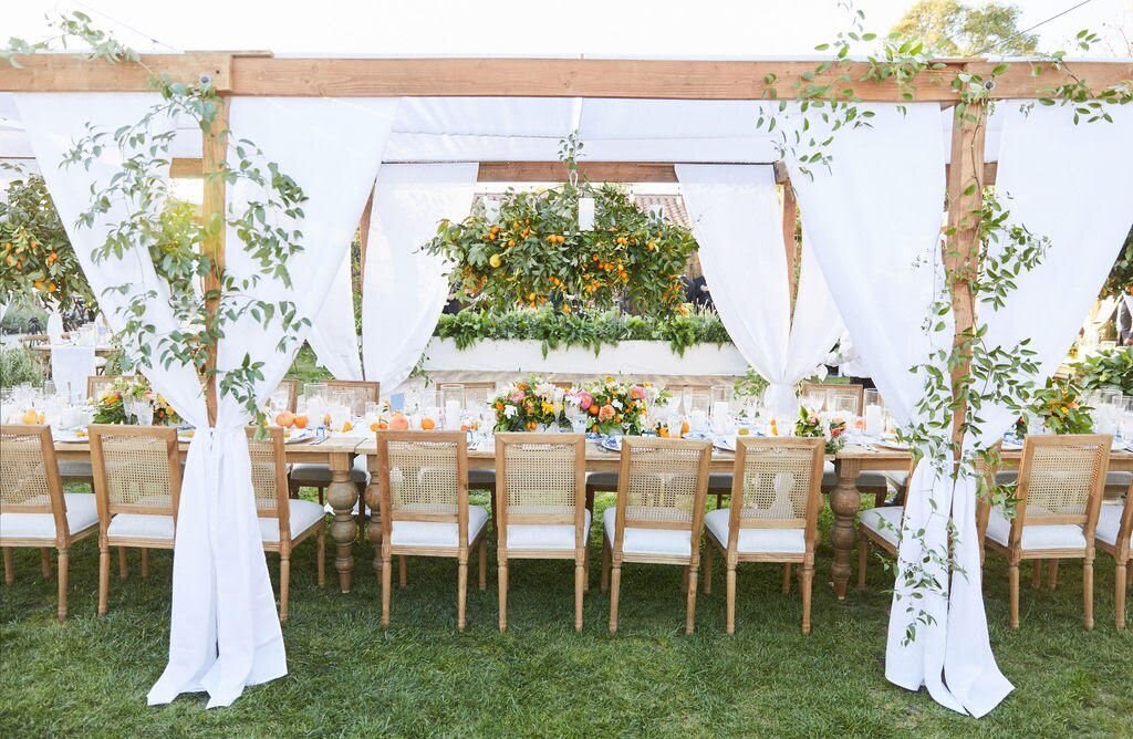 Outdoor wedding reception table on grass, there is a shade frame around the table with flowing white drapes. The space is decorated with small orange fruits and plants.