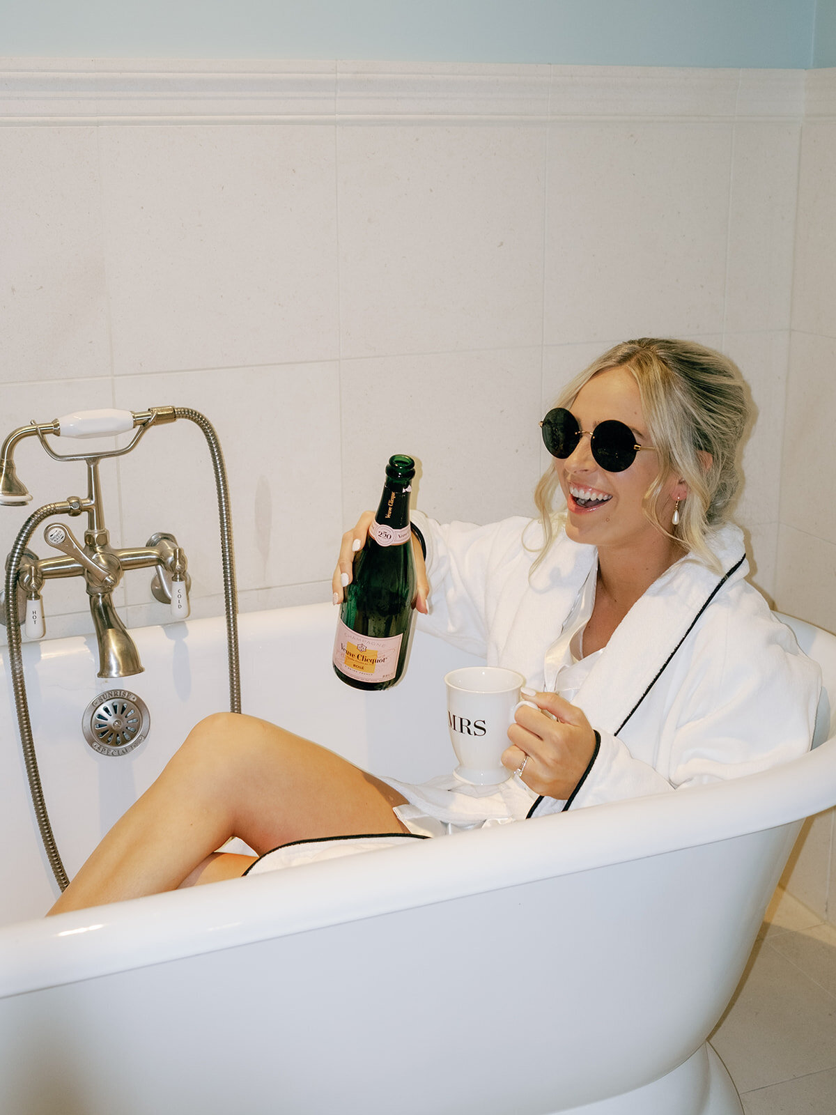 Bride celebrates by ouring champagne and hanging on a bath tub on her wedding morning.