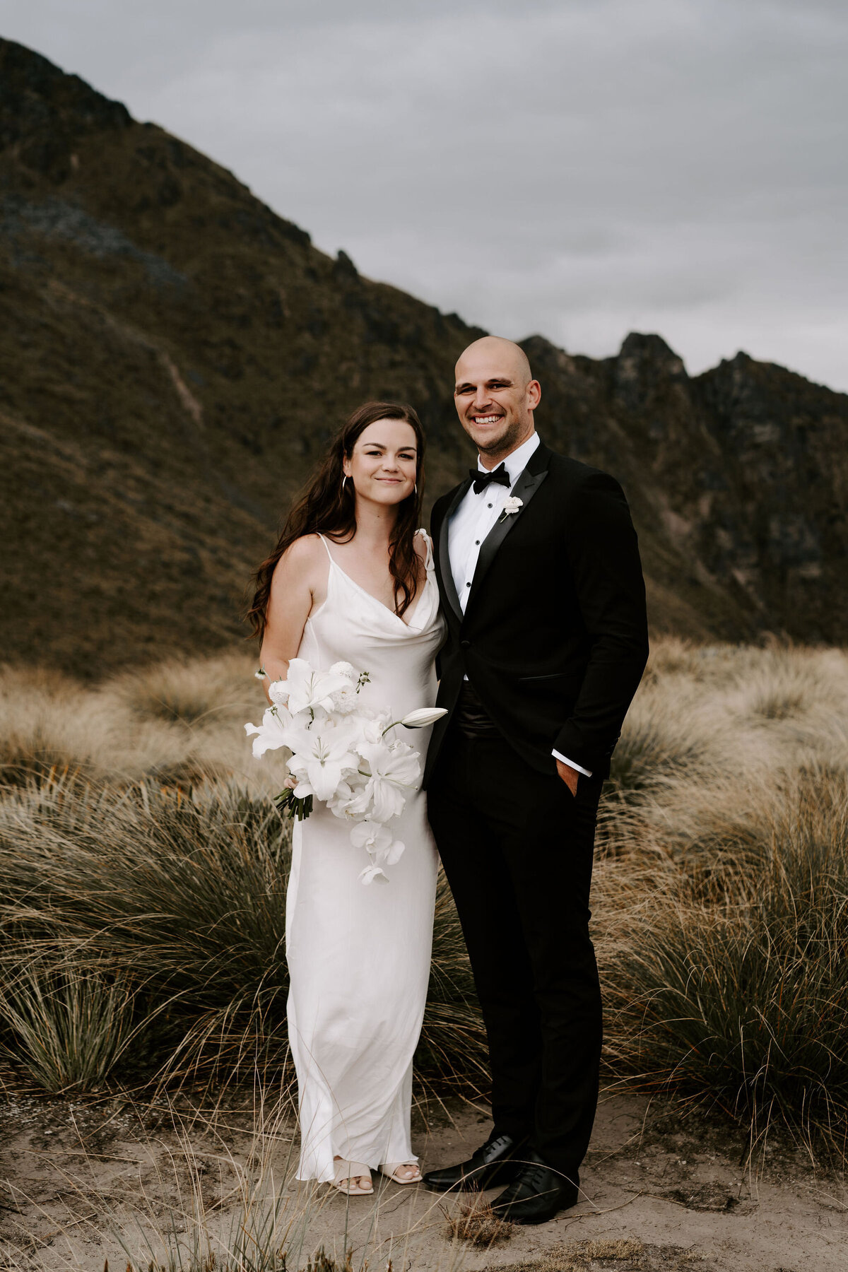 The Lovers Elopement Co - bride and groom pose in long grass on mountain with lake below, heli wedding