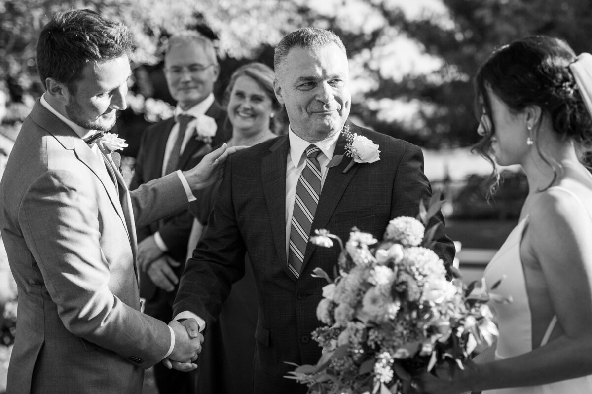 a black and white photo of a father shaking hands with the groom as he and his daughter approach the wedding altar.  Taken by Ottawa wedding photographer JEMMAN Photography