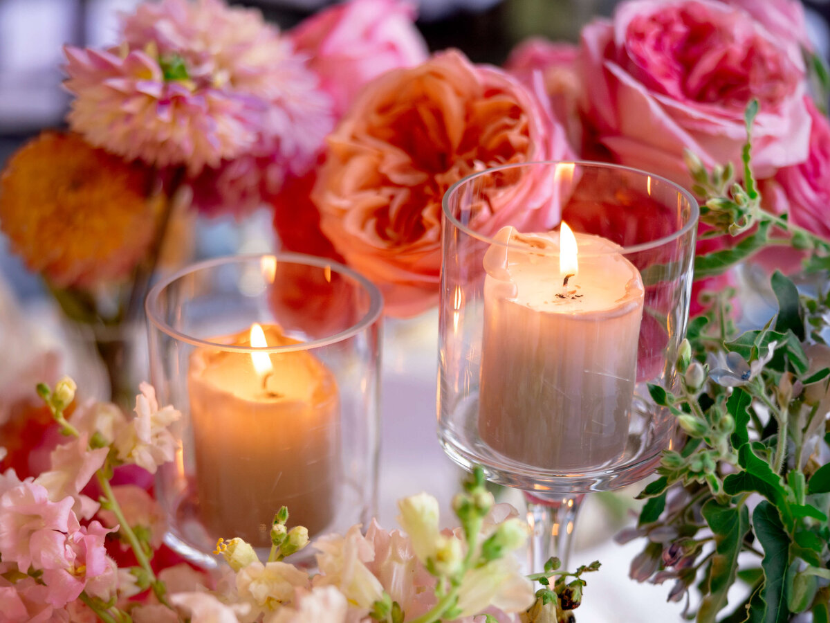 A close-up of lit candles in glass holders surrounded by pink, orange, and peach flowers. Soft lighting enhances the delicate petals and warm glow of the candles, creating a serene and inviting atmosphere.