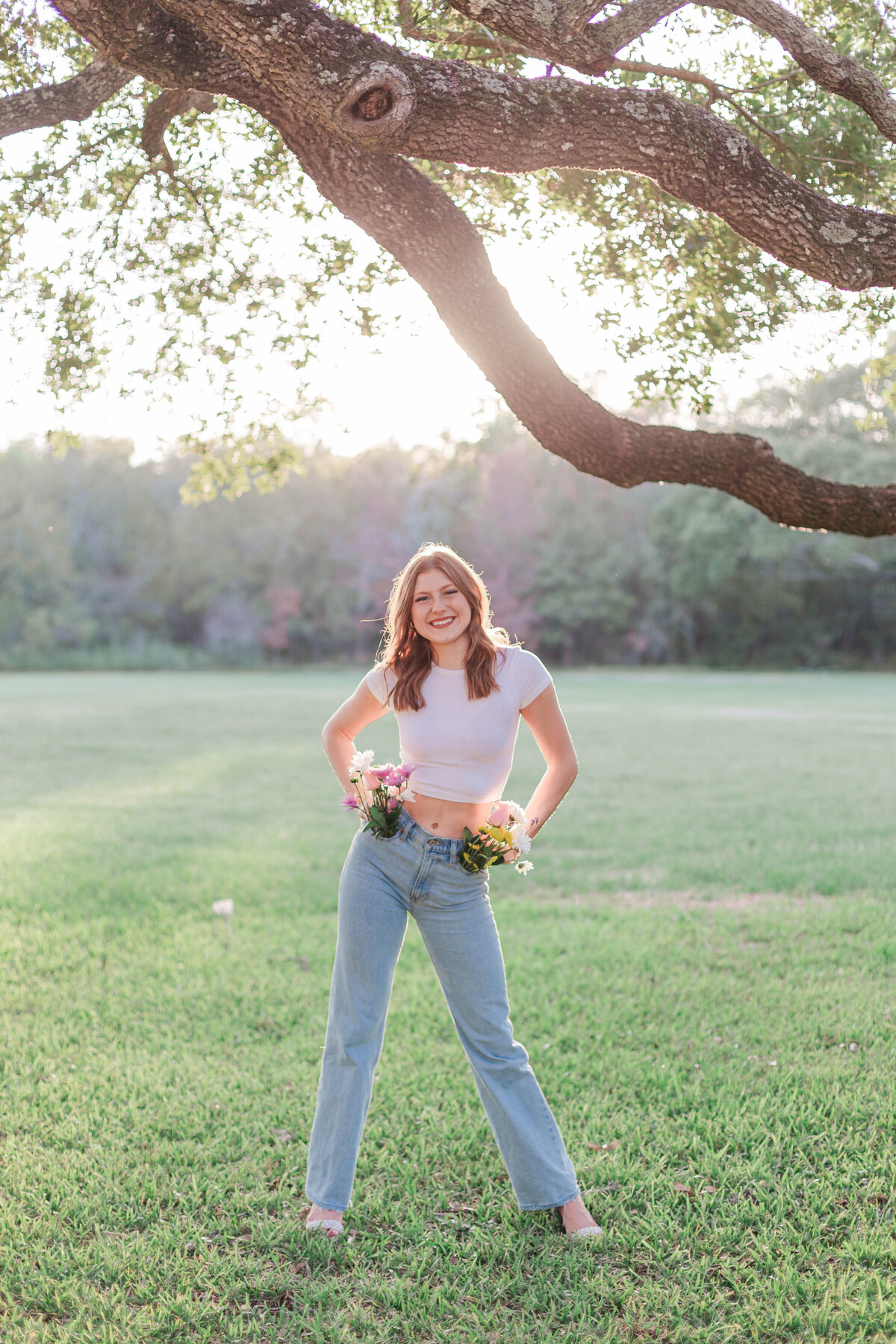A senior girl stands with her hands on her hips wearing a white shirt and jeans with flowers coming out of her pockets in a field in League City Texas
