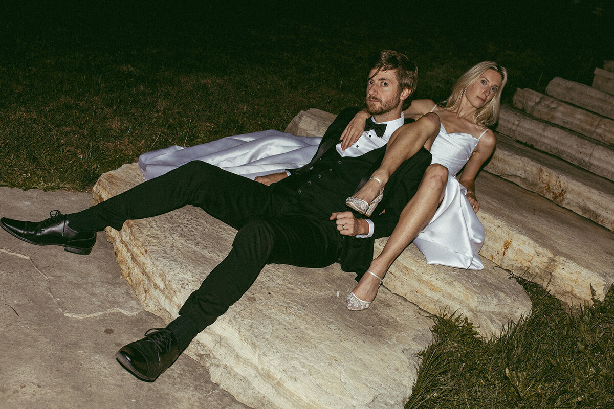 A man in a tuxedo and a woman in a white dress sit casually on stone steps at night, with the man leaning back and looking at the camera during an event in Davenport.