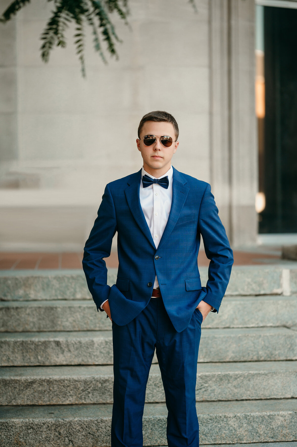 A male student from Waukesha North High School stands on the courthouse steps wearing a navy suit, bowtie and aviator sunglasses during his senior portrait session.