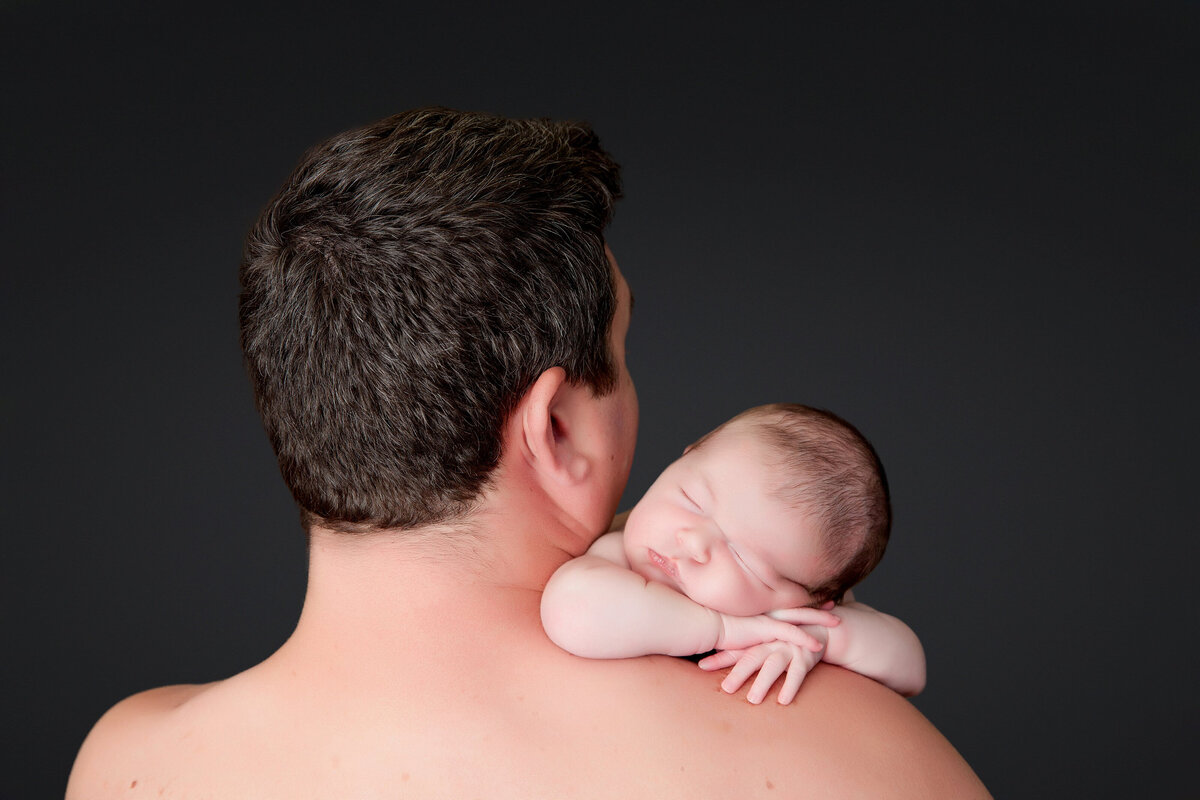 A baby sleeps peacefully, resting on an adult's bare shoulder against a dark background. The Jacksonville newborn photographer captures this tender moment as the adult, turned away, reveals a head of short, dark hair. The baby's arms are gently draped over the adult's shoulder, embodying serenity.