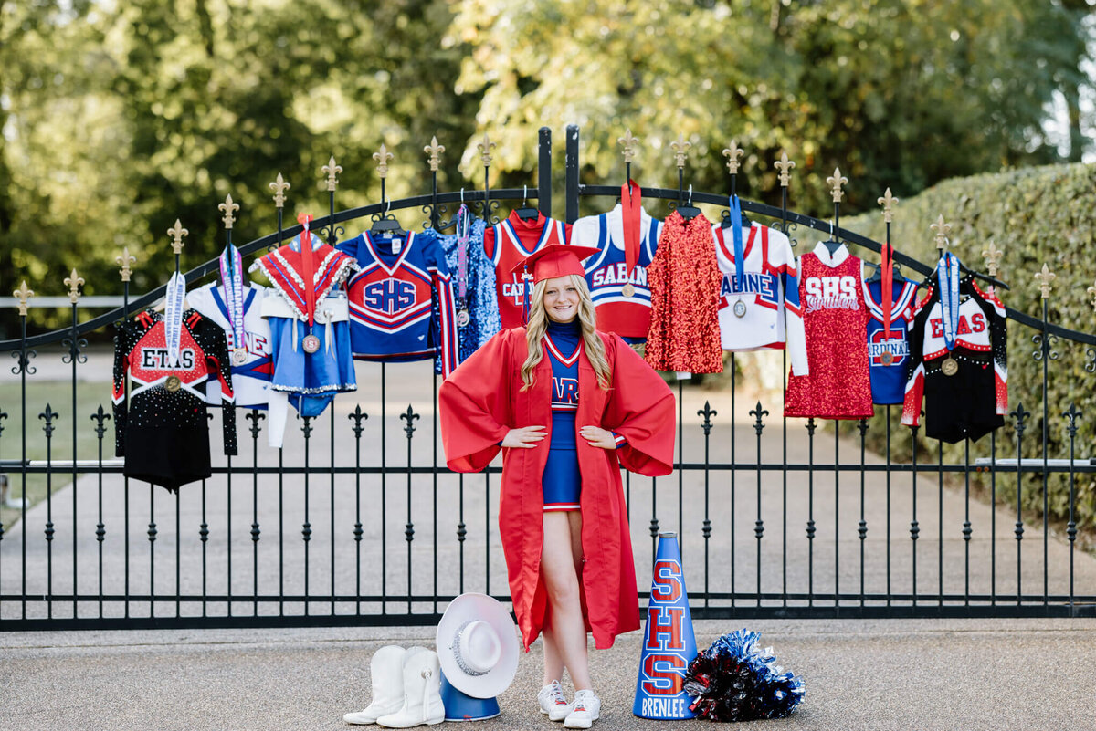 Sabine High senior girl in cheerleading uniform and cap and gown standing in front of gate displaying all cheer and dance medals and past uniforms