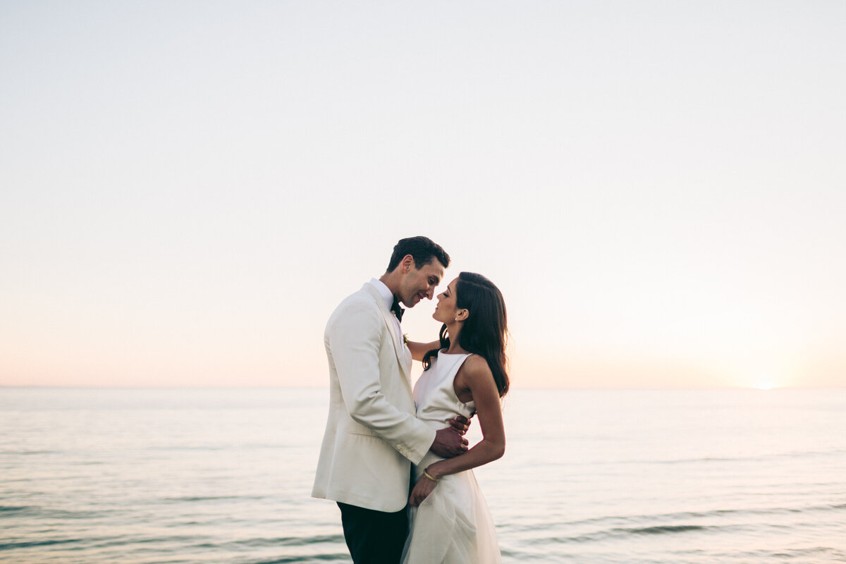 A photograph in color of Sarah and Erik on their wedding day at Dos Pueblos Orchid Farm in Santa Barbara, California. The portrait is taken just after sunset so it has an overall pastel glow and soft light. The ocean and sky are behind the bride and groom and they blend into one another, separated by a slightly orange horizon. The ocean is calm with gentle waves. They are facing each other with his arm on her waist and they are closing in for a kiss. He is in a tuxedo with a white jacket and black pants and she is in a white satin wedding dress with a high neckline and narrow straps. Wedding photography by Stacie McChesney/Vitae Weddings.