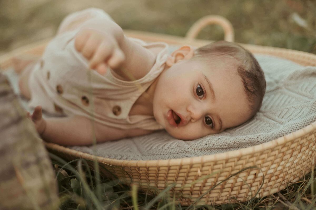 A curious baby in a linen romper lying on a basket reaching for long blades of green grass.