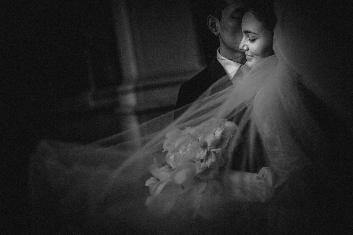 A groom kissing a brides cheek as her veil flows around her.