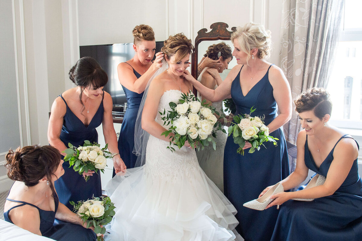 bridesmaids in navy dresses holding bouquets help an elegant bride get ready for her Chateaul Laurier wedding in the Prime Minister's Suite.  Captured by Ottawa wedding photographer JEMMAN Photography