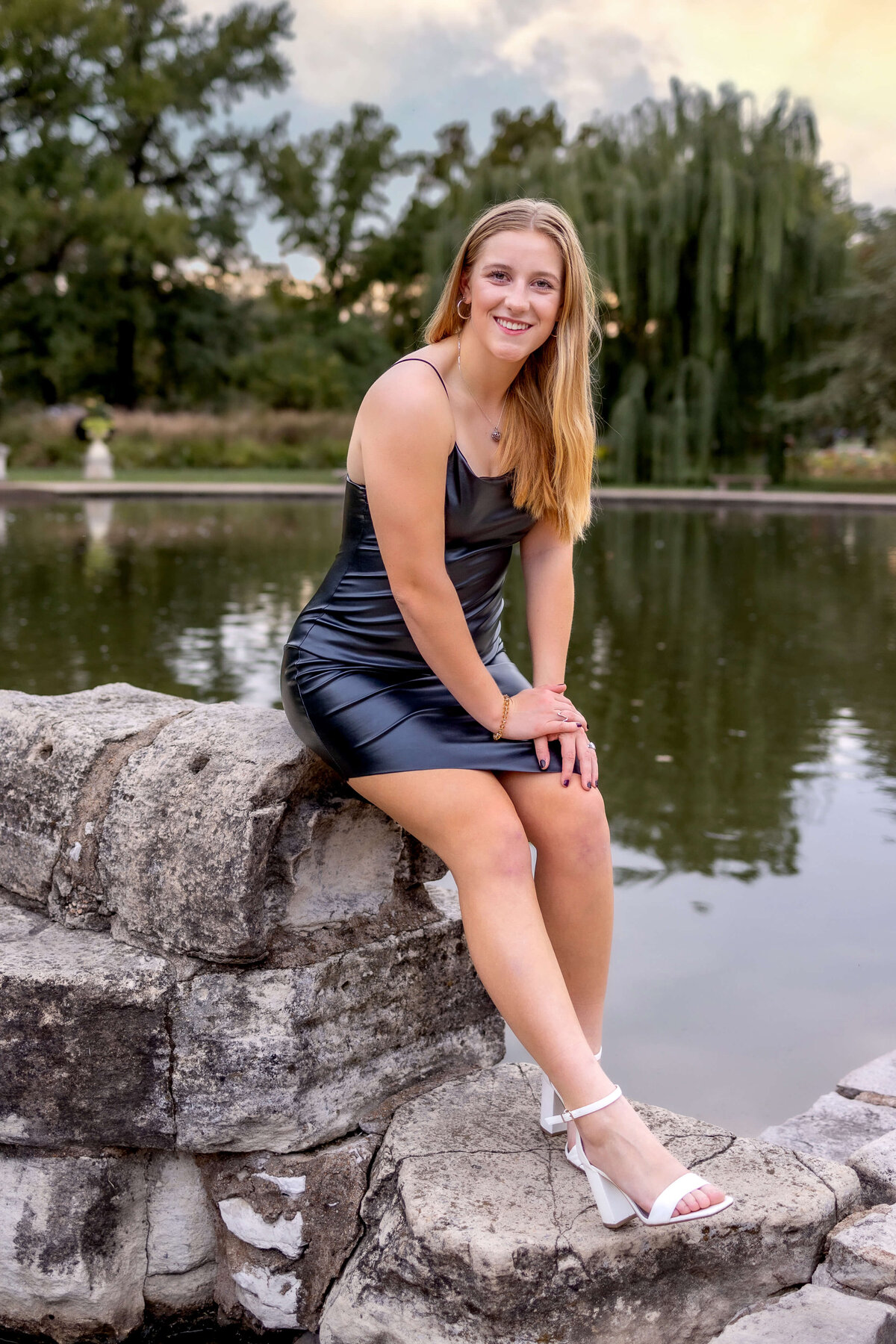 A long blonde haired  girl is sitting and leaned forward onto her knees next to a stone sculpture and a big pond in Tower Grove Park.