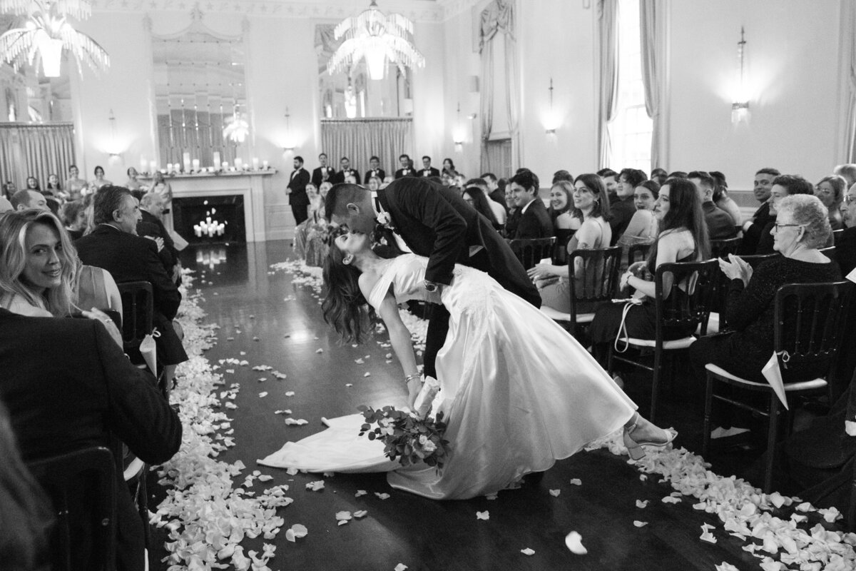 Black and white photo of a groom dipping his bride for a kiss at the end of the aisle after ceremony New York wedding photographer Milestone Images