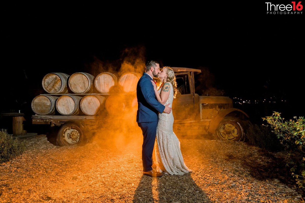 Newly married couple share a kiss standing in front of a vintage rustic truck carrying old wine barrels with a brownish mist behind them