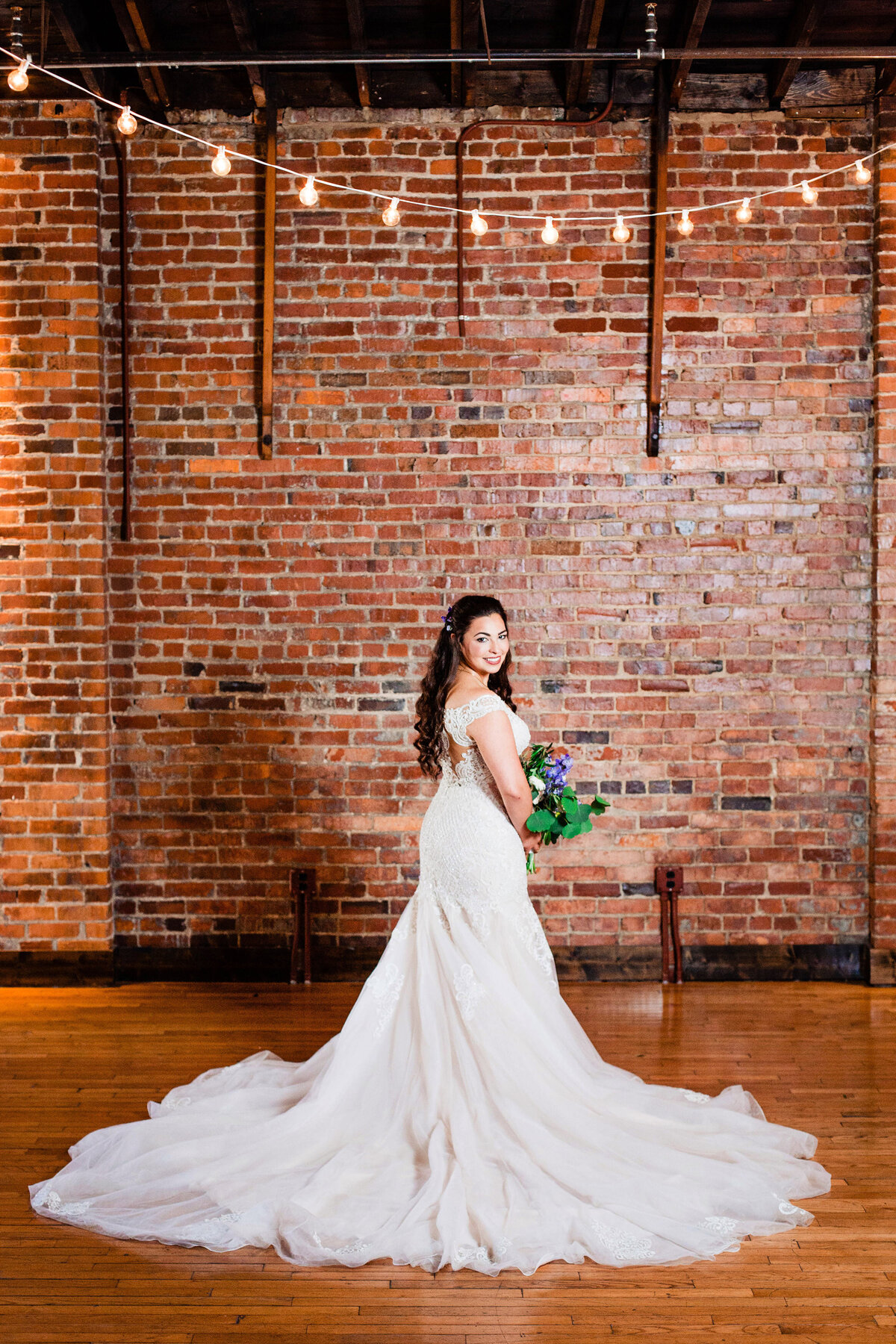 Brides train fanned out and she's looking over her shoulder smiling at the camera, brick wall behind her