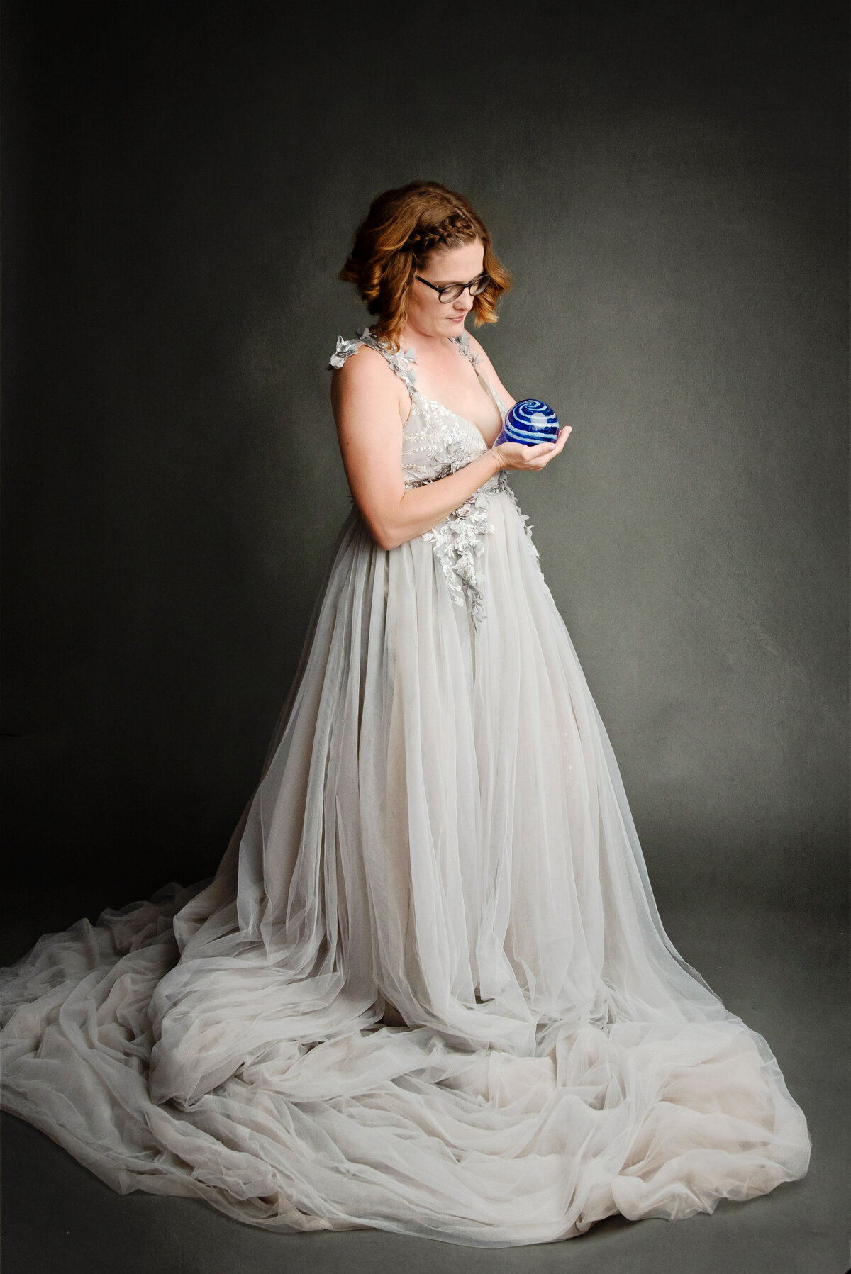 st-louis-motherhood-photographer-mom-in-flowy-silver-gown-holding-blue-and-white-glass-ball-against-gray-background