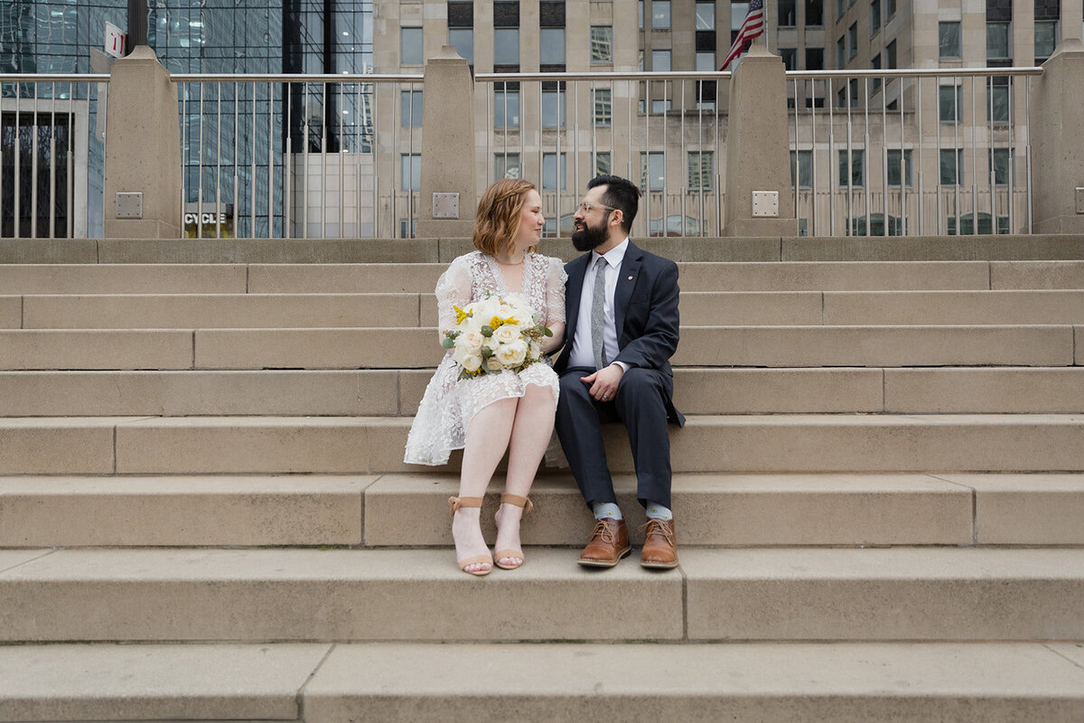 Wedding couple sitting on steps along the Chicago Riverwalk. They're looking at each other.
