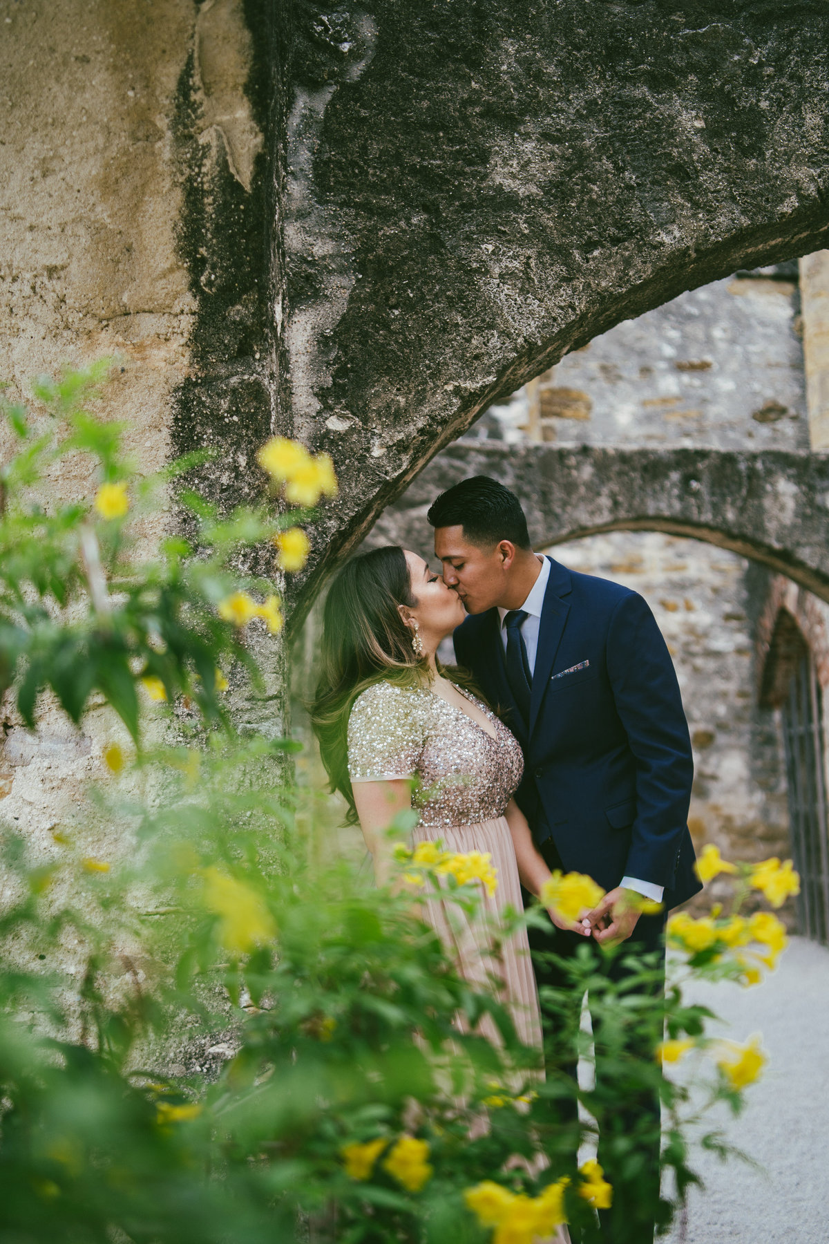 Engaged man and woman kissing behind yellow flowers and under an arch at Mission San Jose in San Antonio, Texas.