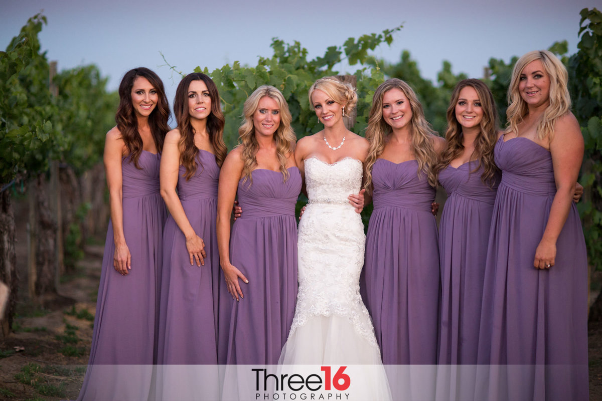 Bride poses with her bridesmaids all dressed in lavender while standing in the orchard