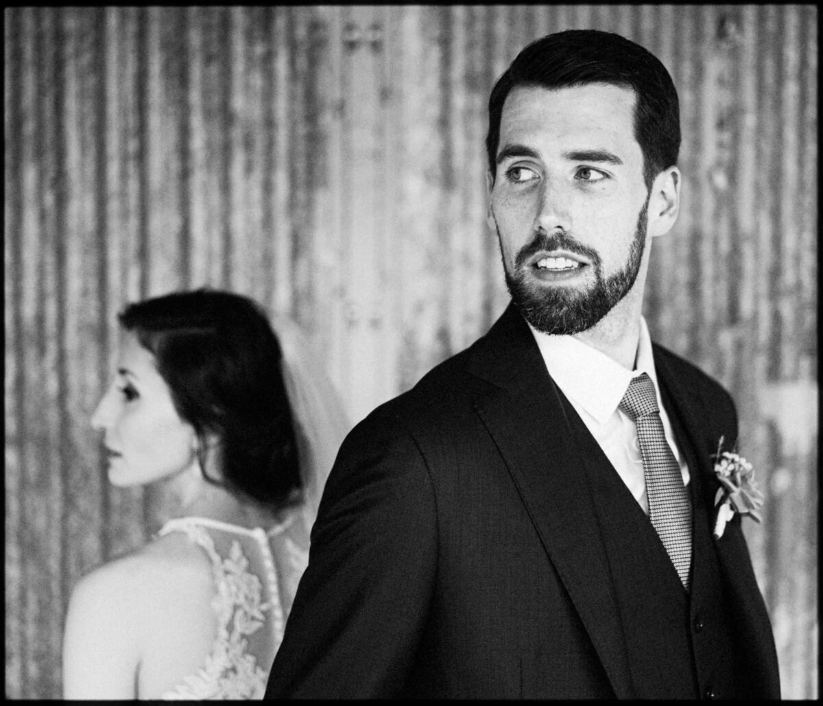 Black and white photo of a bride and groom, focused on the groom's expression as he looks to the side with the bride in the background
