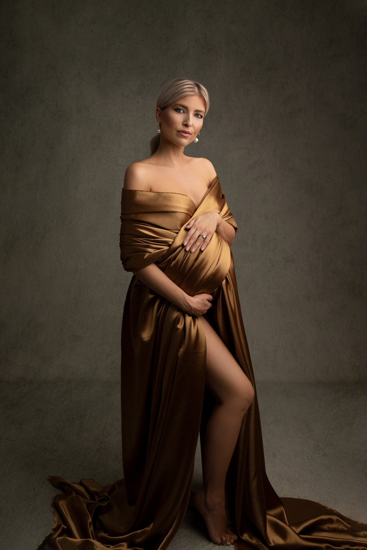 Woman poses for fine art maternity photos with New Jersey's best maternity photographer Katie Marshall. Woman is in a ruched, gold silk wrap  that puddles on the floor. She is angled slightly away from the camera with her front knee bent in the slit of the fabric. One hand is under her bump, the other over her bump. She is looking at the camera with a closed-mouth smile.