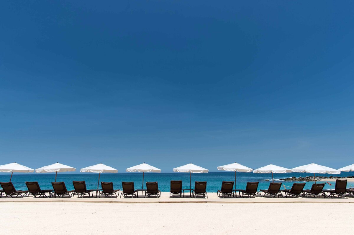 A line of chairs and umbrellas are shot on a beach at noon with horizon and blue sky