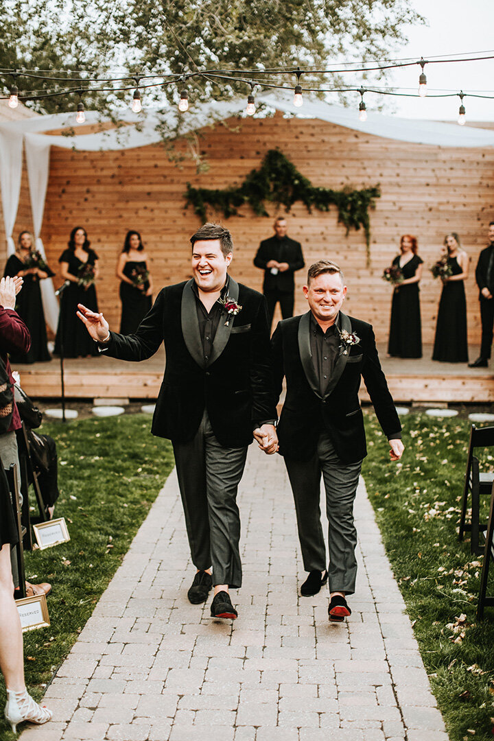 Two grooms wearing black tuxedos walk down the aisle holding hands smiling and waving at their wedding.