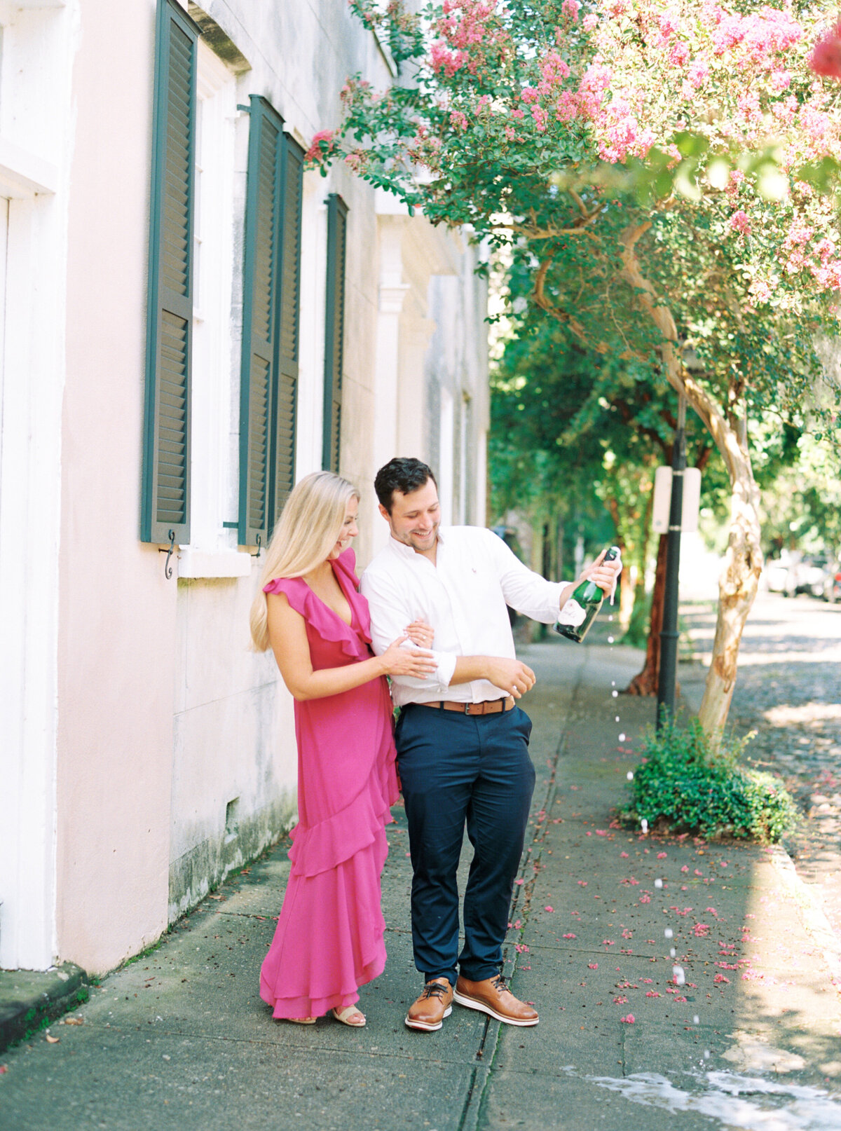 Engagement session with couple popping champagne on the sidewalk in downtown Charleston with pink blooms. Girl in bold pink dress.