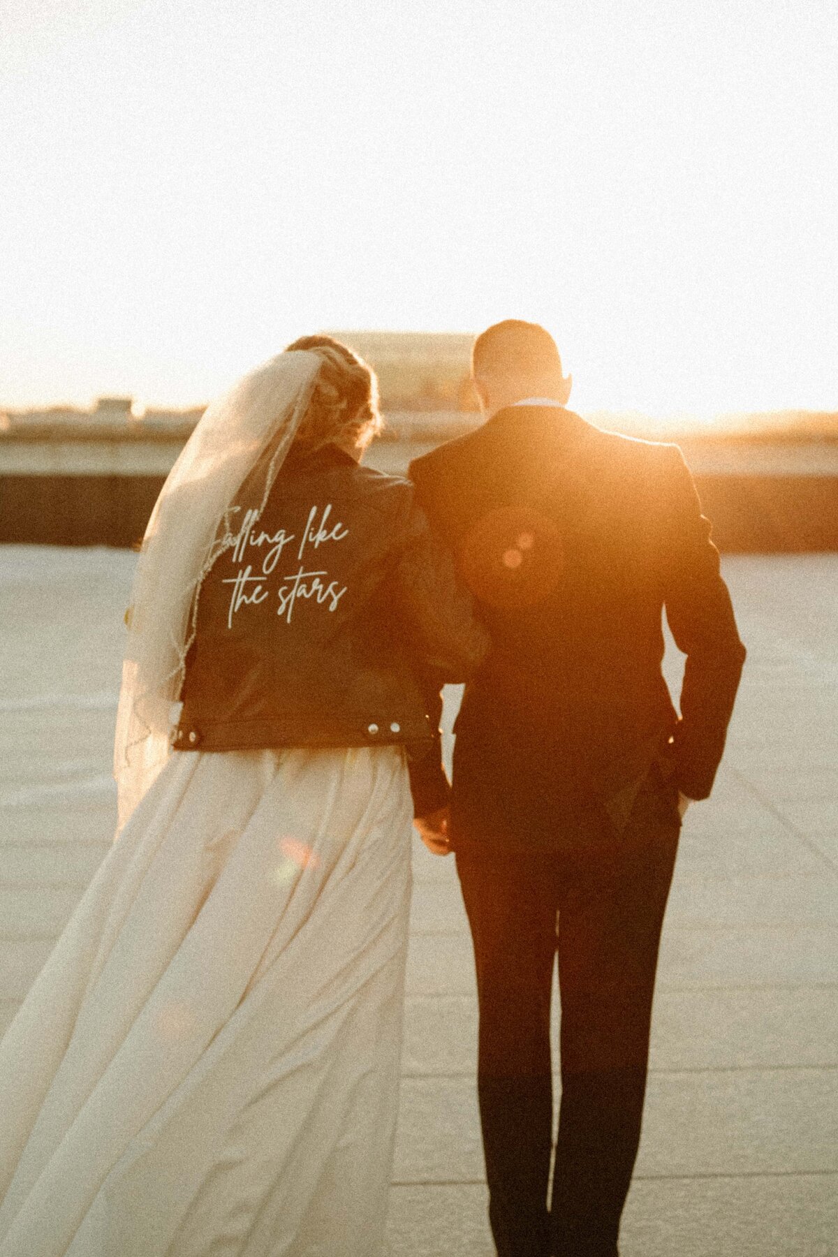 A bride in a white dress and a groom in a suit walk hand in hand during an Iowa sunset, with the bride's jacket embroidered with "bring me the stars.