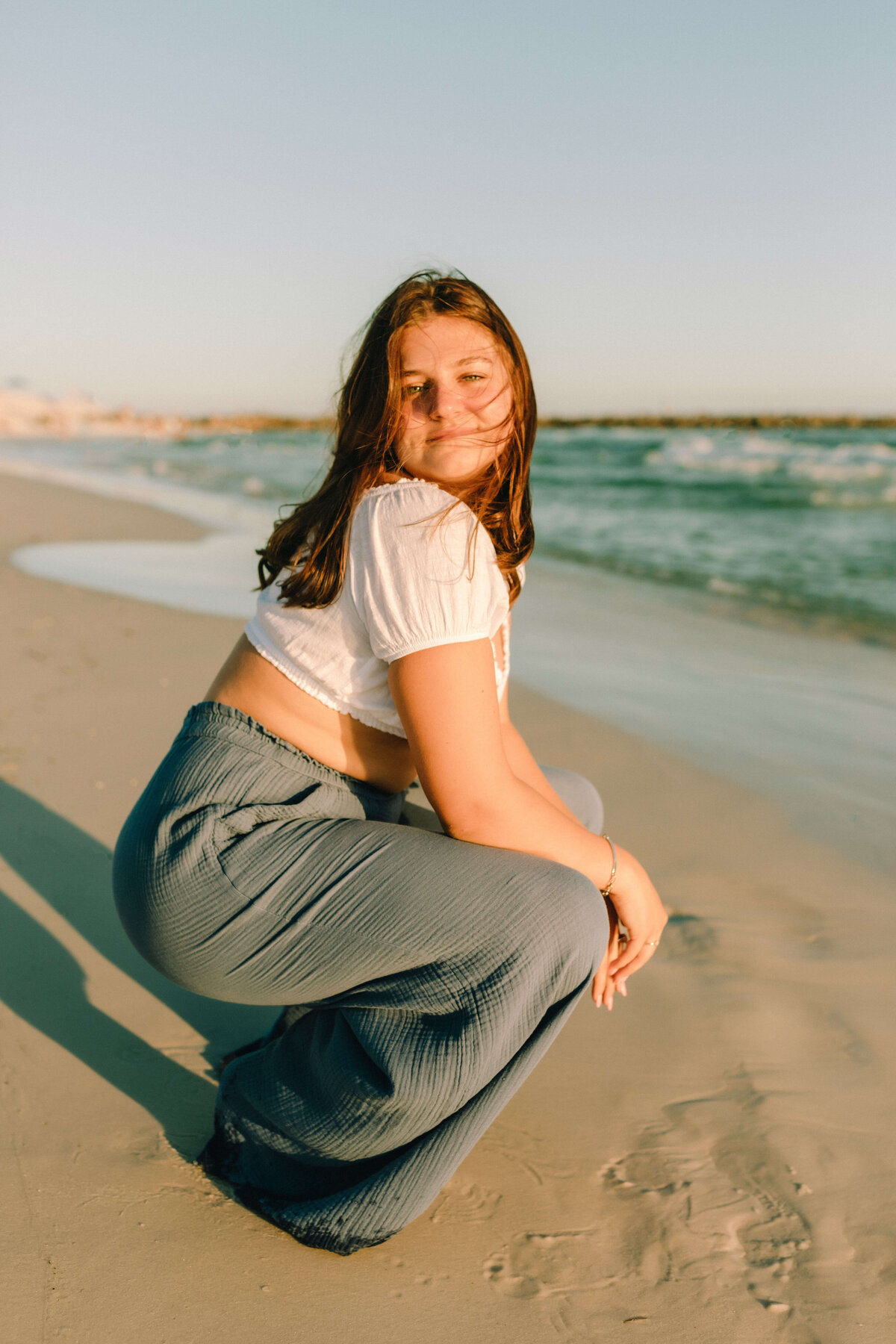 brunette senior girl in white crop top & blue loose pants crouches in sand - portrait taken by panama city fl photographer brittney stanley