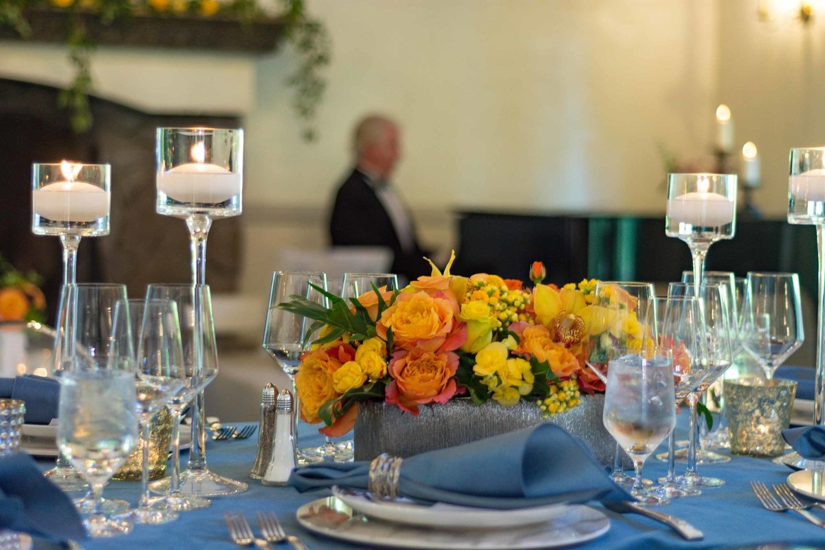 orange flower centerpiece on blue linen with floating candles in stemmed glass candle holders