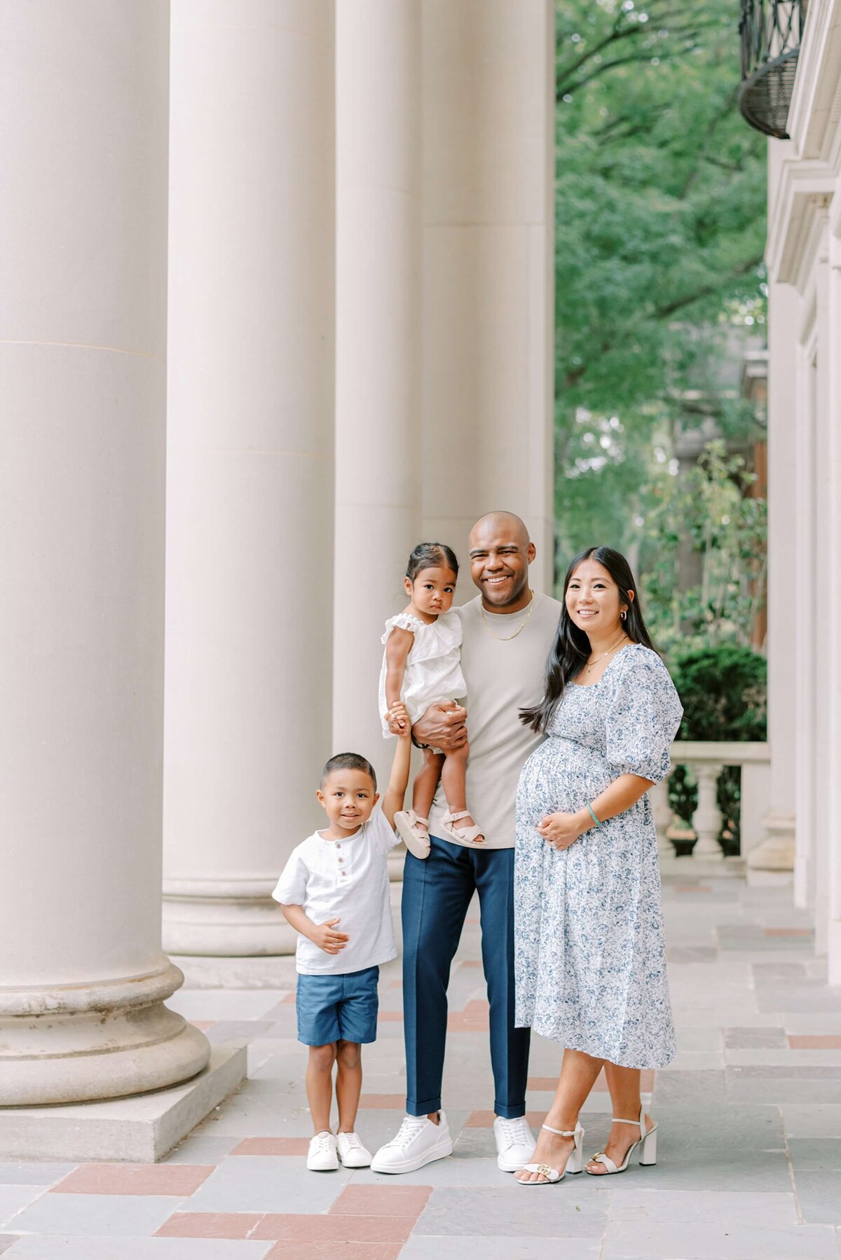 A family of four pose together for a portrait while mom cradles her baby bump next to tall columns outside of  a stately building on Davidson College's campus.