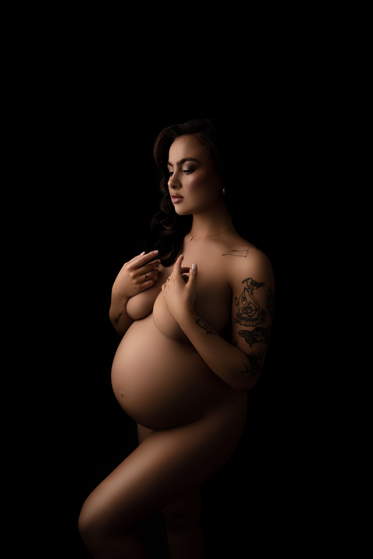 In this captivating maternity photo, expertly crafted by Katie Marshall, Philadelphia Main Line's premier maternity photographer, a bare-skinned woman stands in an alluring profile pose. Her hands artfully conceal her breasts, while one knee is gracefully bent, showcasing her unique tattoos. The play of dark shadows and subtle highlights masterfully accentuates her silhouette. This striking image celebrates the beauty of motherhood and exemplifies the artistry of Katie Marshall, a renowned maternity photographer in the Philadelphia Main Line area.
