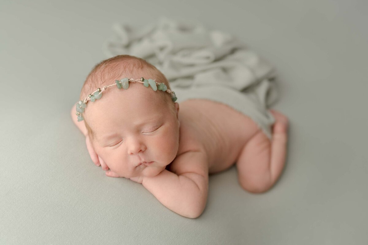 Newborn baby girl laying on tummy with chin resting on hands laying on sage blanket with matching jewel headband and swaddle