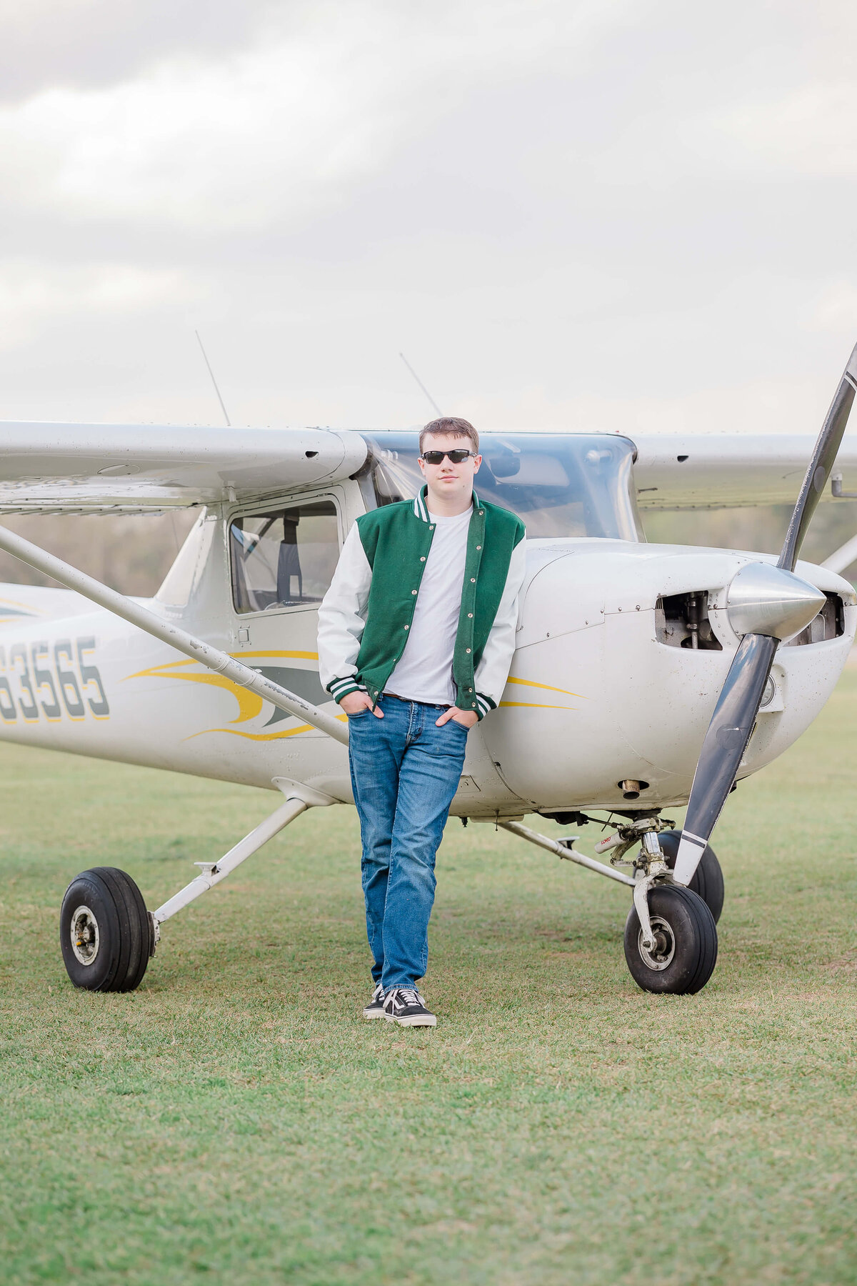 A person in sunglasses and a green varsity jacket leans against the nose of a small white airplane with a propeller. They are standing on grass, with overcast skies in the background.