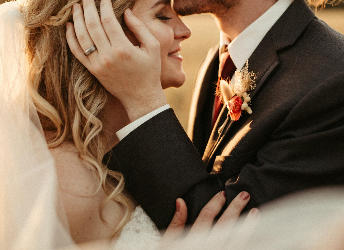 Close up of groom holding bride's face and kissing her forehead as she smiles and holds his arm at sunset