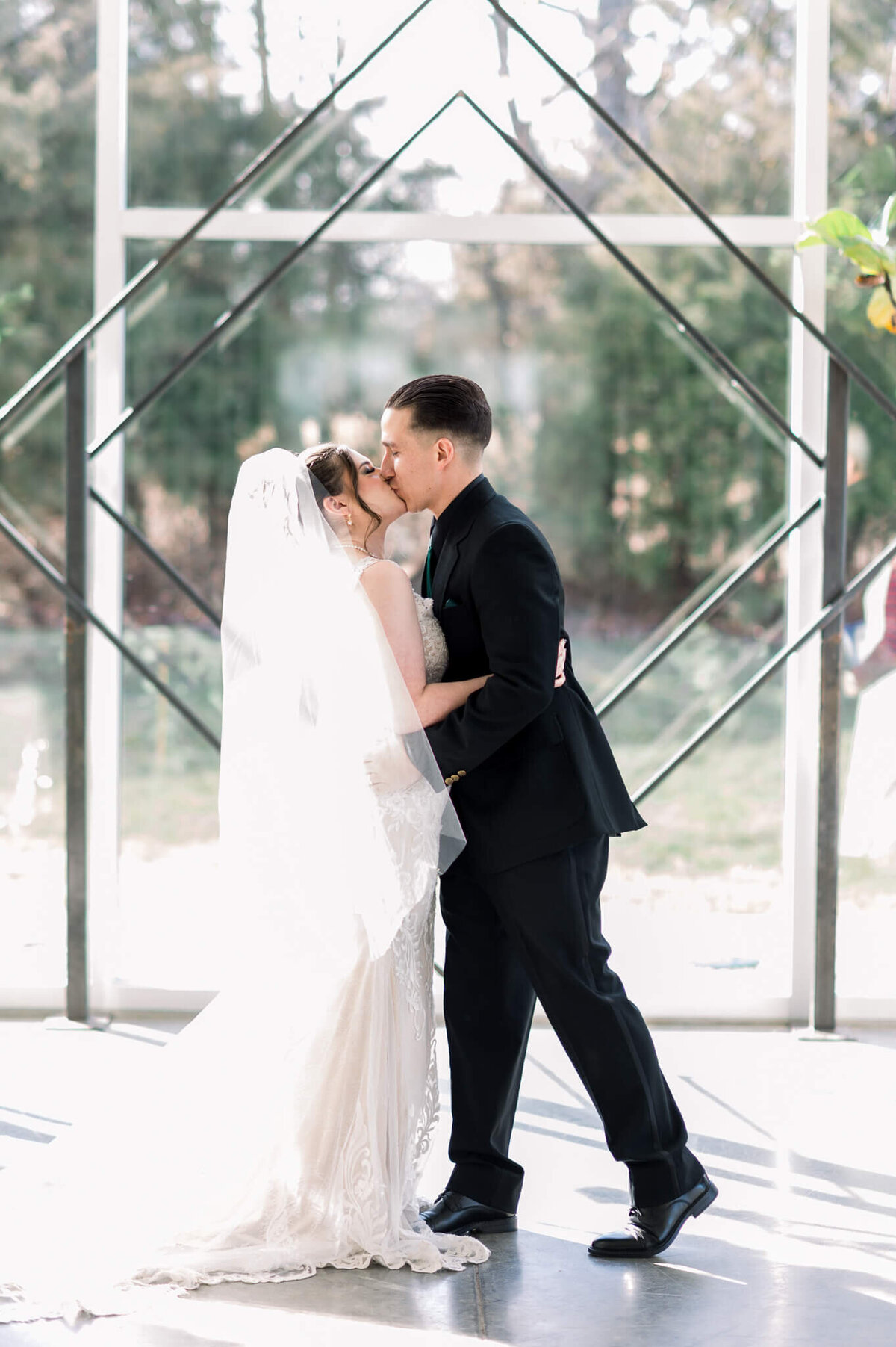 couple shares first kiss as husband and wife, bride wears white dress with hip length veil, groom wears black suit, wedding arbor in background is two diamonds