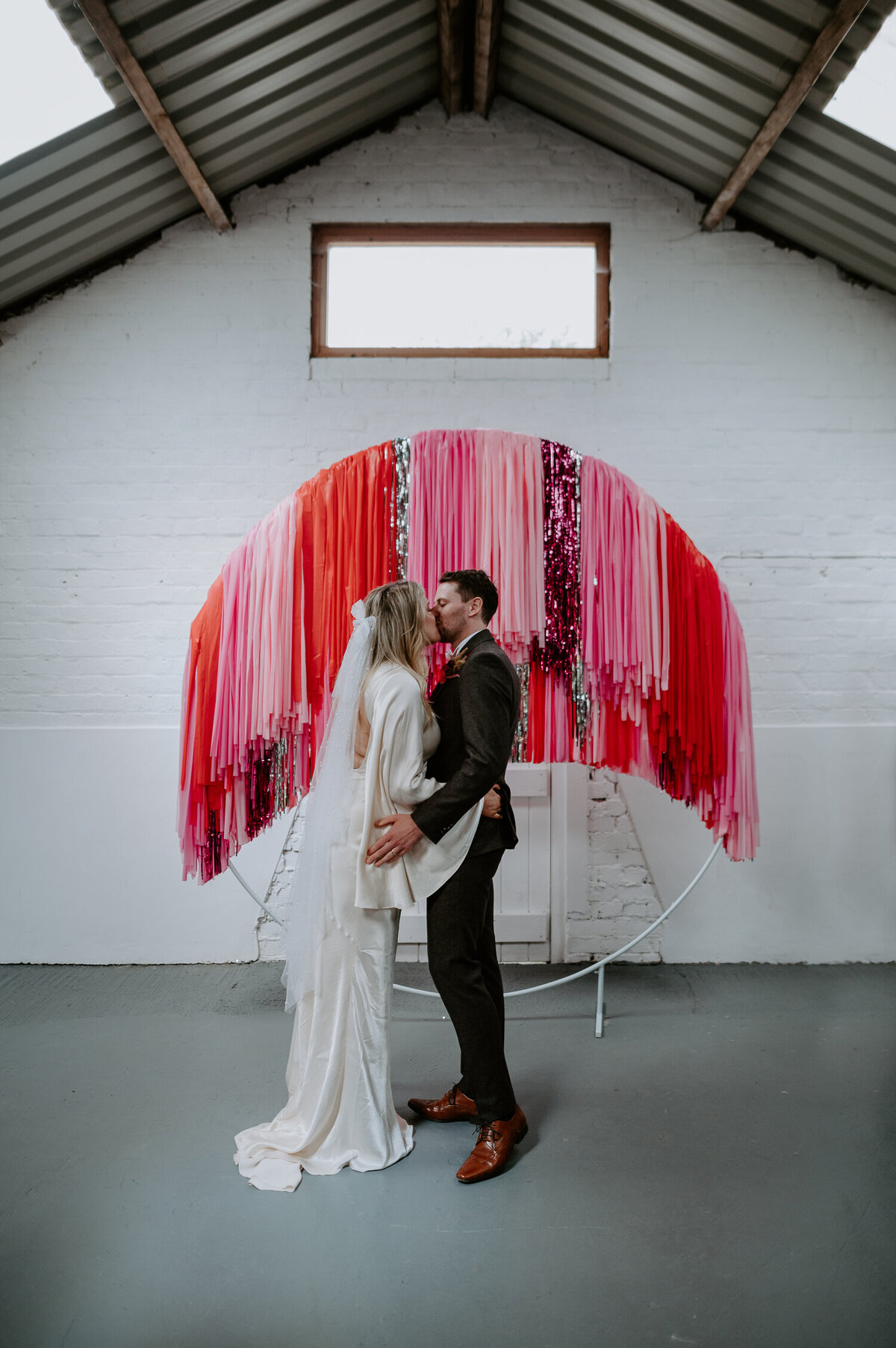 A couple share a first kiss at the White Syke Fields. They are stood infront of a streamer backdrop which was a DIY project the bride made.