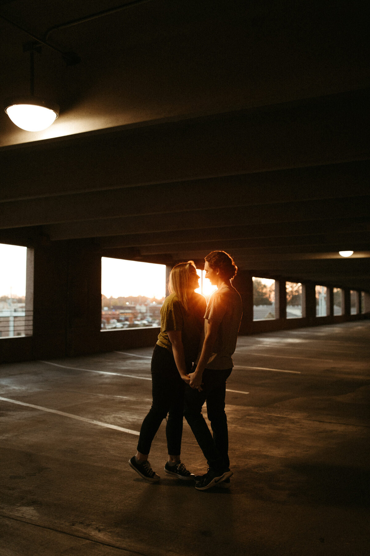 A couple is standing next to each other holding hands inside the lower level of a parking garage as the sun sets behind them.