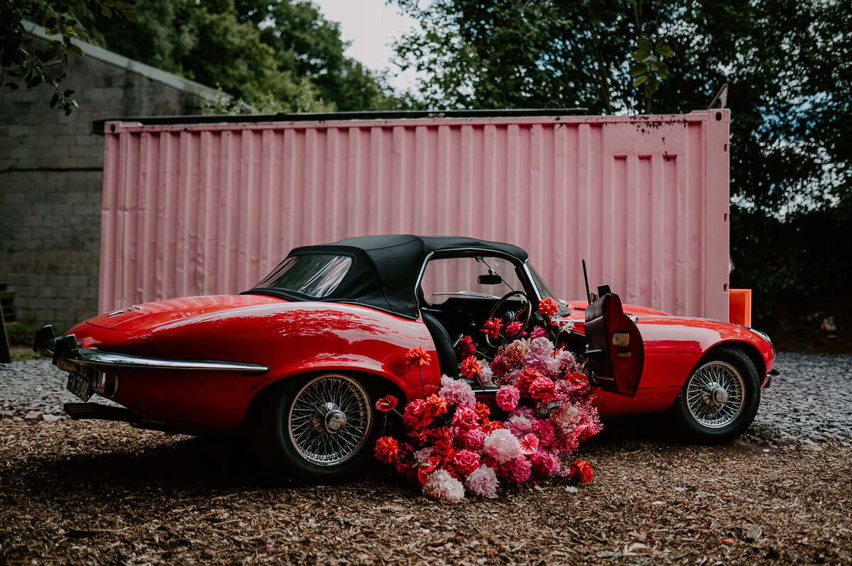 A huge pink and red floral instalment inside of a classic red soft top Jaguar, in the background is a pink shipping container which was painted for a DIY wedding.
