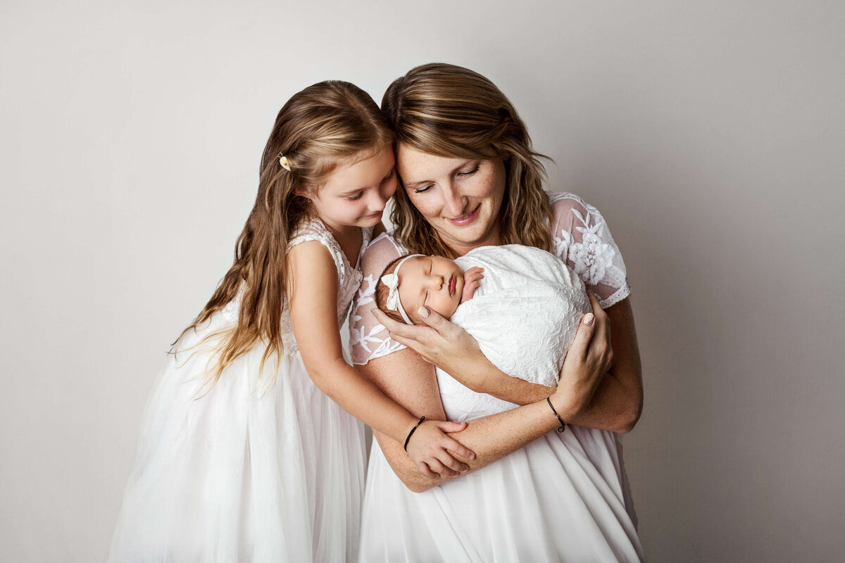 mom and daughter wearing pretty white dresses holding a newborn baby in a white wrap on a white backdrop