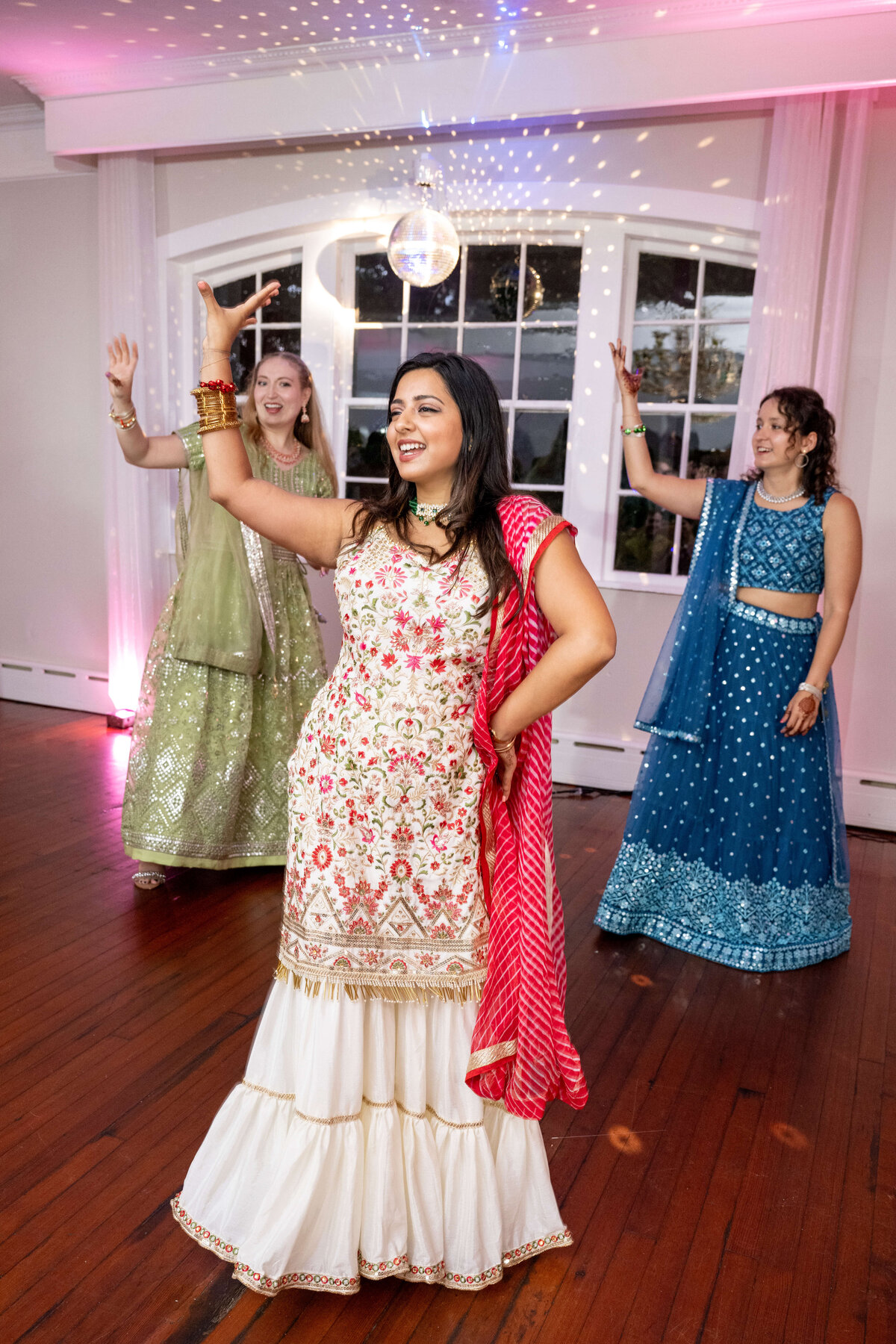 Three women dance joyfully under a disco ball in a room with large windows. They wear colorful, traditional South Asian outfits in shades of white, pink, green, and blue. The wooden floor adds warmth to the setting.