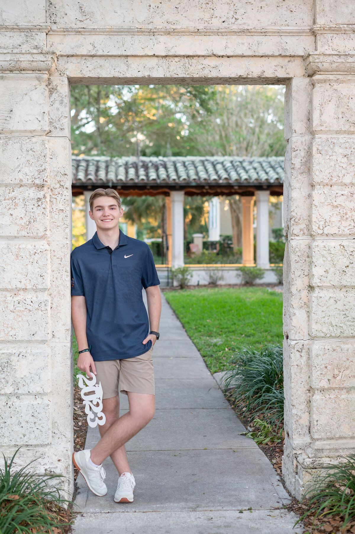 High school senior boy holding "2023" wooden sign leaning against stone doorway.