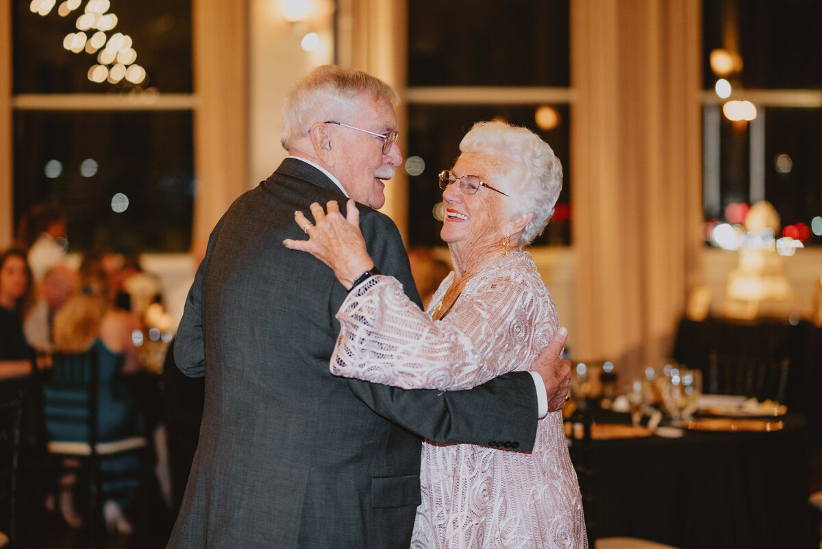 An elderly couple joyfully dances in an elegant setting, smiling warmly at each other. Dim lighting and softly glowing chandeliers create a romantic ambiance.