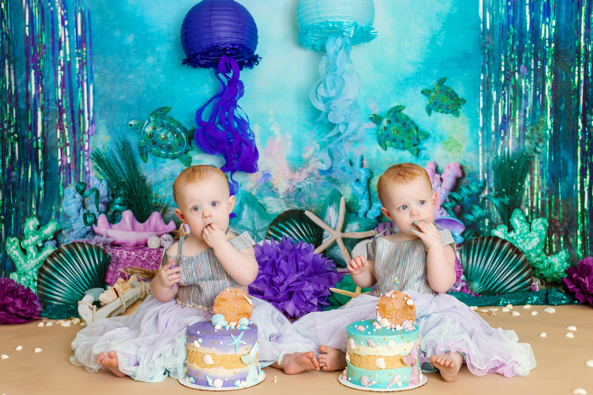 Cake smash photographer, twin girls sit eating cake with a ocean backdrop