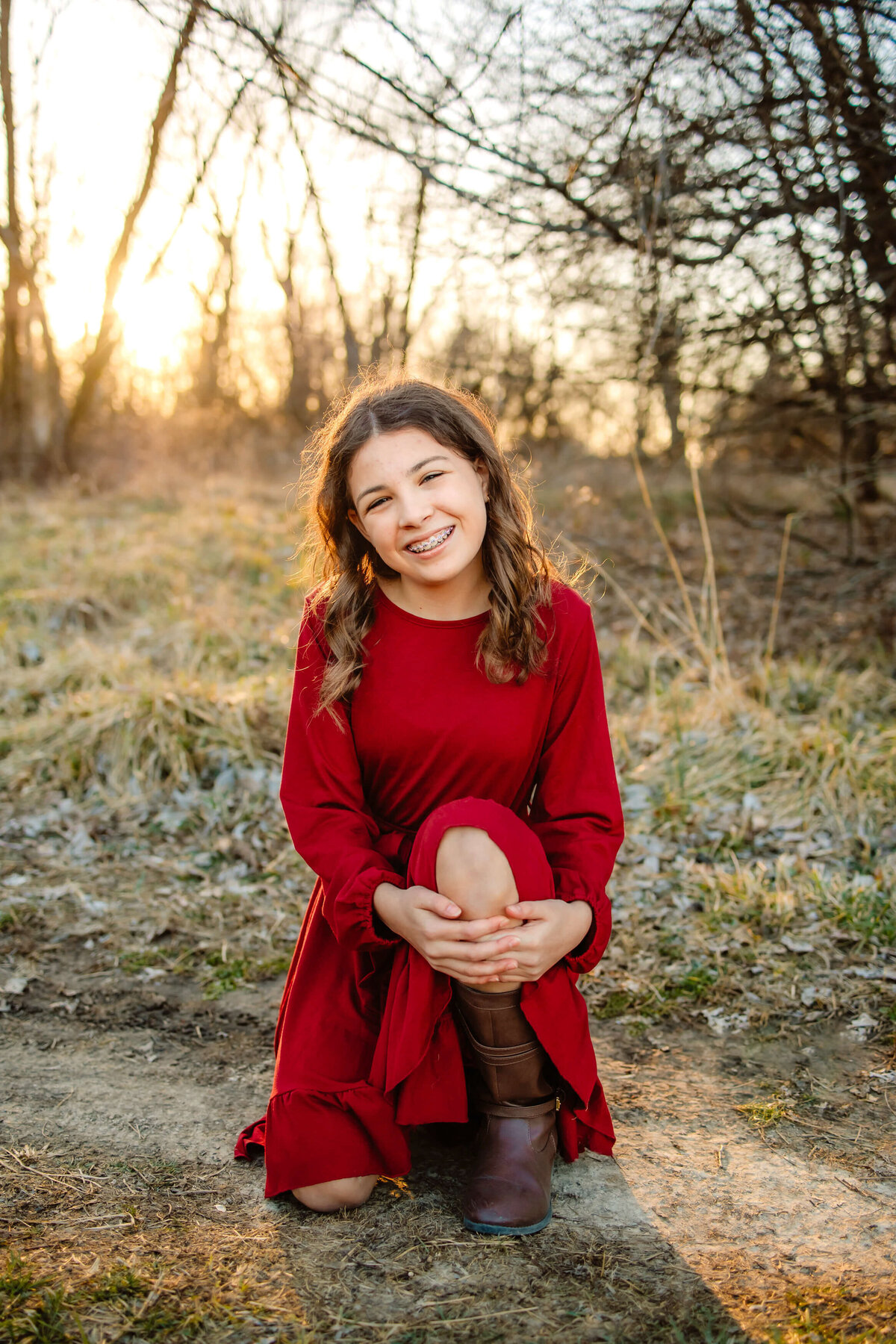 beautiful young lady in a long red dress sitting down on a dirt path  of a field at sunset