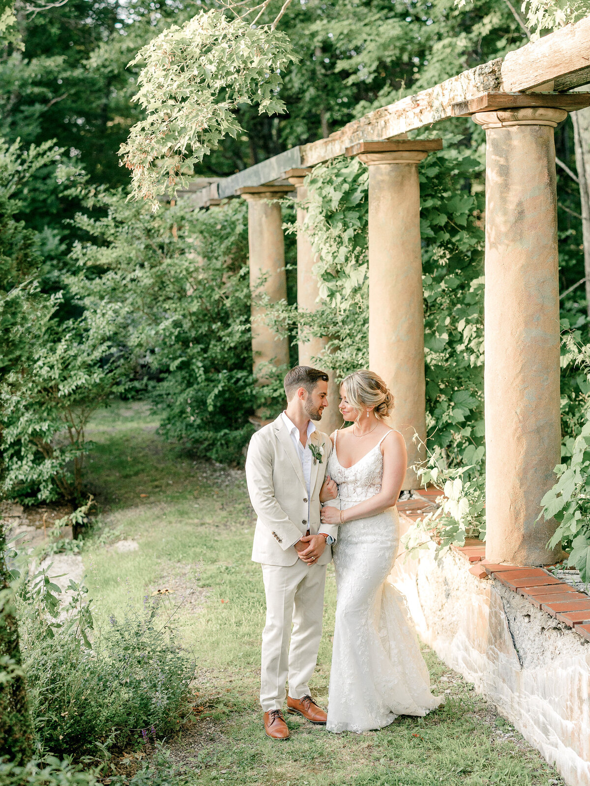 Bride and groom in a tan suit standing in a garden with old columns and greenery