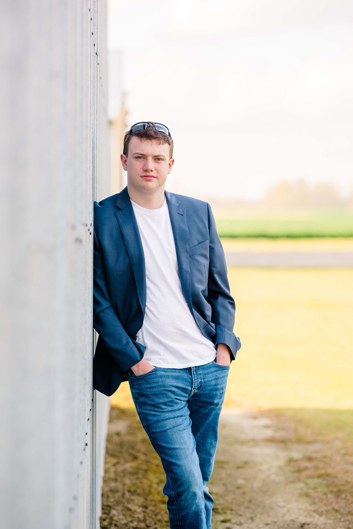 A person wearing a blue blazer, white shirt, and jeans leans against a wall outdoors on a sunny day in Fayetteville, NC. They have sunglasses resting on their head and their hands in their pockets. The scene captures the essence of senior photography with grass and fields in the background.